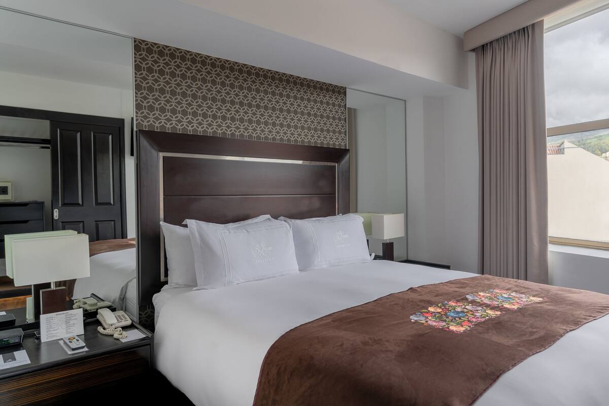 Quarto de hotel com uma cama grande, mesa de cabeceira com telefone e janela com vista para edifícios e árvores.