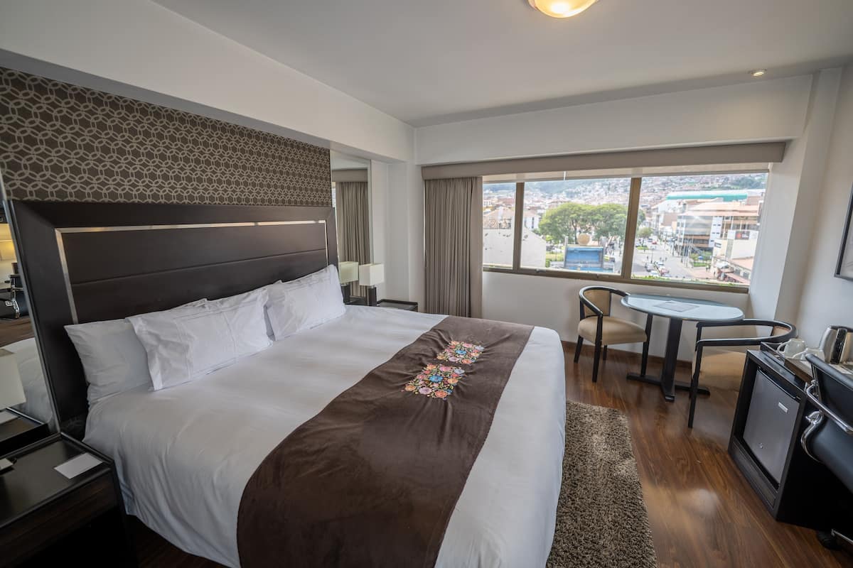Quarto de hotel com uma cama grande, uma escrivaninha, uma cadeira e vista para a cidade pela janela.