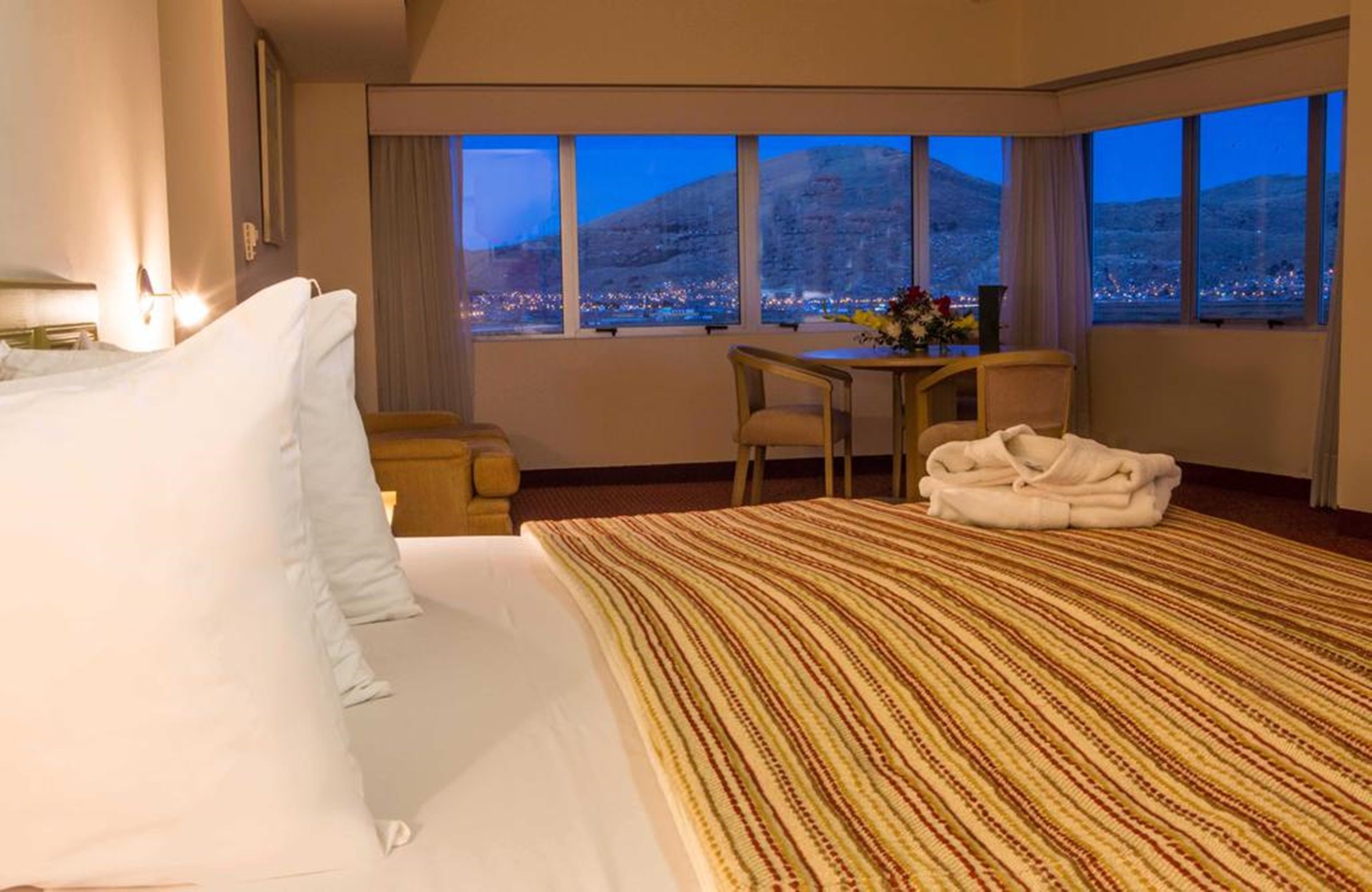 Habitación de hotel con una cama grande, un escritorio y vistas a un paisaje urbano al atardecer.