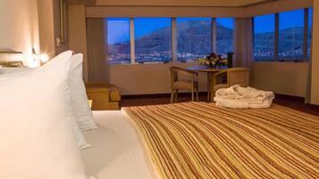 Habitación de hotel con una cama grande, un escritorio y vistas a un paisaje urbano al atardecer.