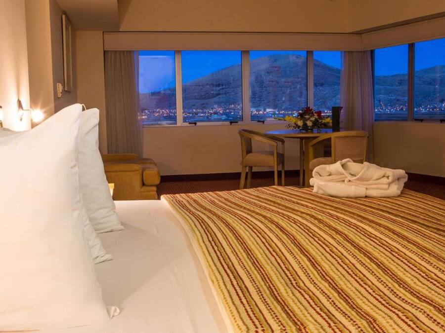 Habitación de hotel con una cama grande, un escritorio y vistas a un paisaje urbano al atardecer.