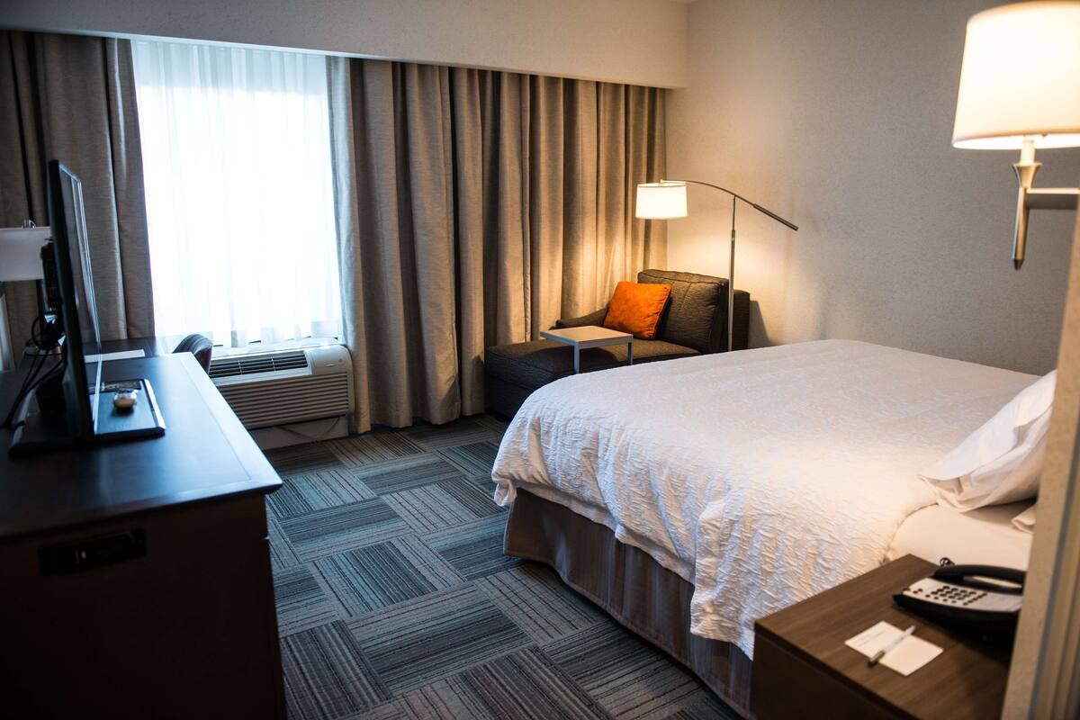 Quarto de hotel com cama, escrivaninha, cadeira, luminária e janela com cortinas.