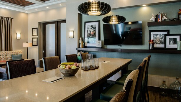 Un comedor moderno con una mesa larga, sillas y un frutero. Hay luces empotradas en la pared y un televisor de pantalla plana sobre la mesa.