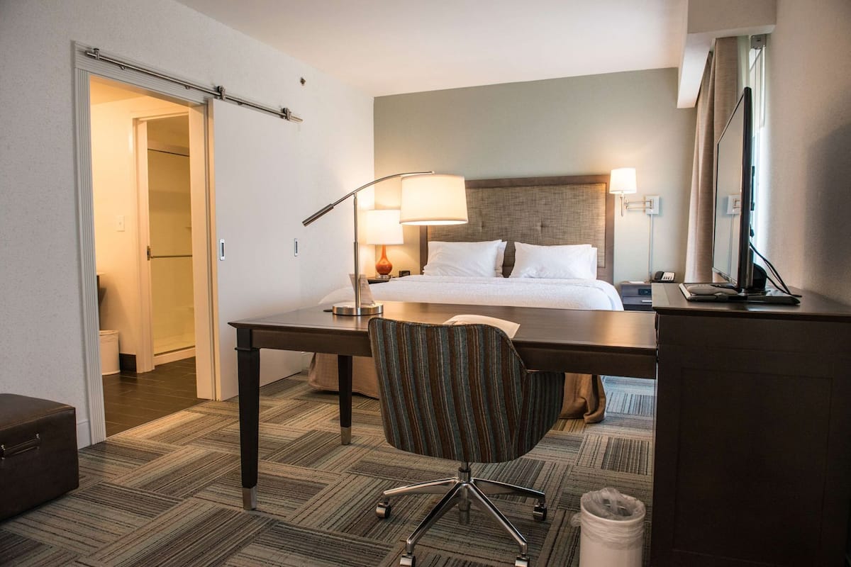 Quarto de hotel com cama, escrivaninha, cadeira, luminária e espelho.