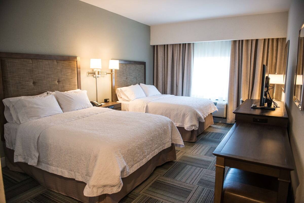 Quarto de hotel com duas camas, uma escrivaninha, uma luminária e uma televisão.