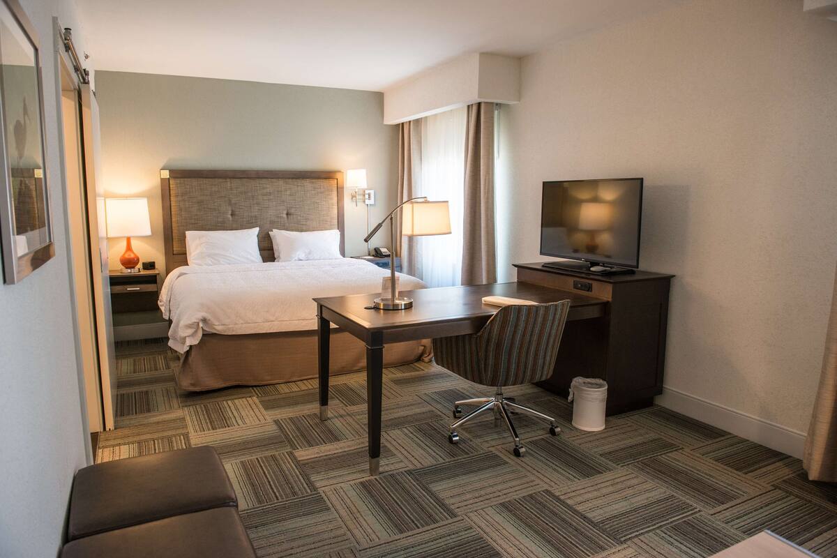 Quarto de hotel com cama, escrivaninha, cadeira, televisão e lumin ária.