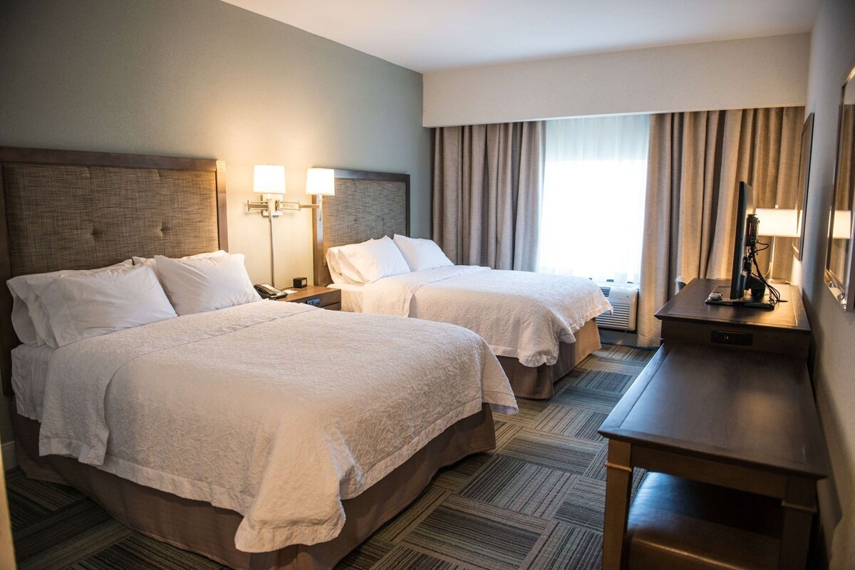 A hotel room with two beds, a headboard, bedside lamps, a desk, and a window with curtains.