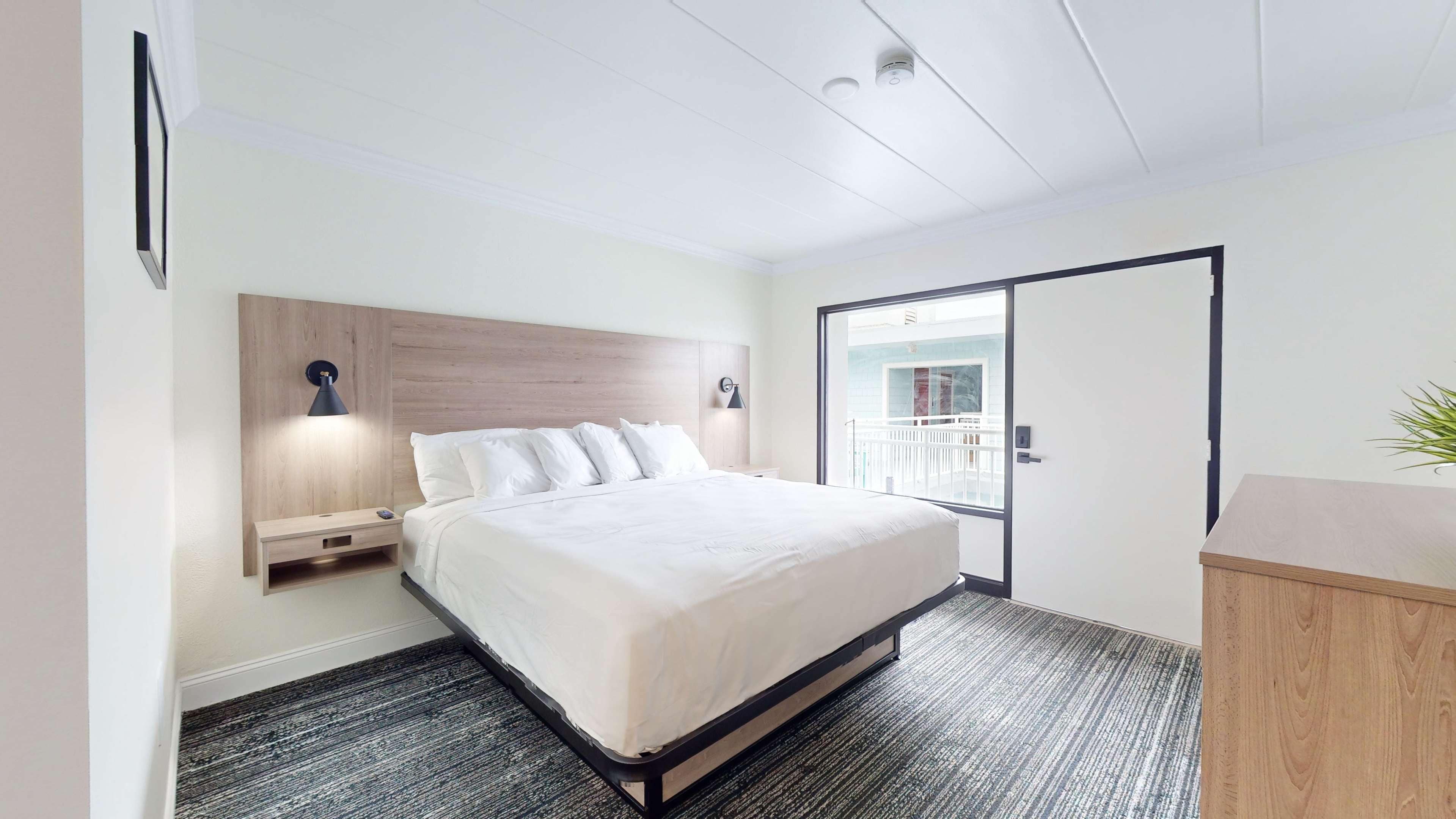 Une chambre d’hôtel moderne avec un grand lit, des lampes de chevet, une tête de lit en bois, une petite table de chevet, un bureau et une vue sur un balcon.