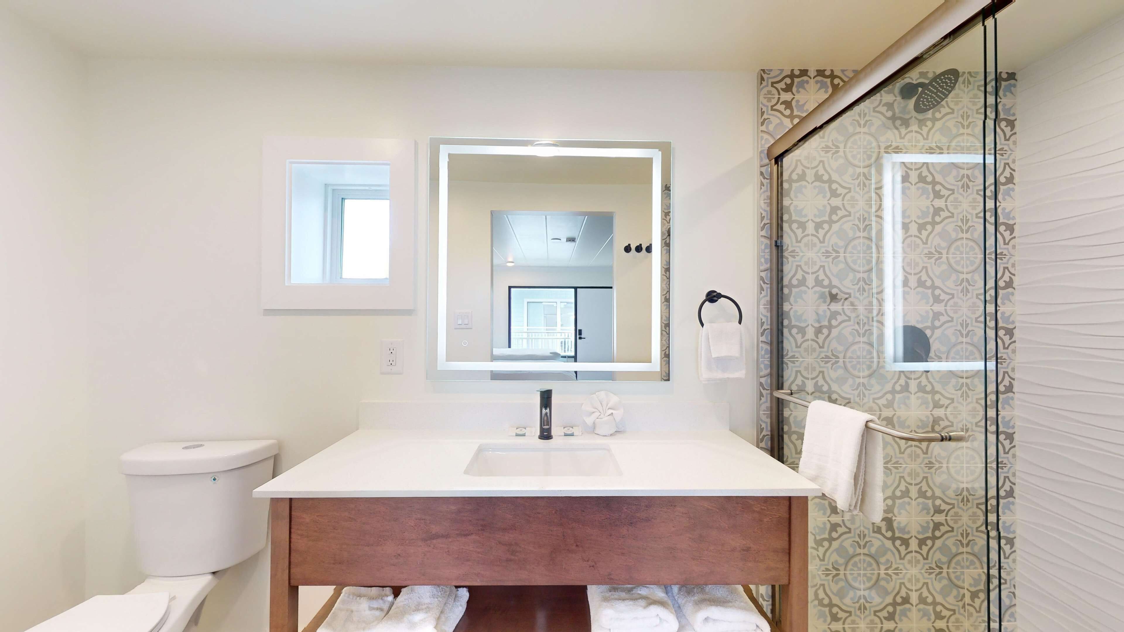 A modern bathroom with a white vanity, a large mirror, a shower with patterned tiles, and a view of the room through the mirror.
