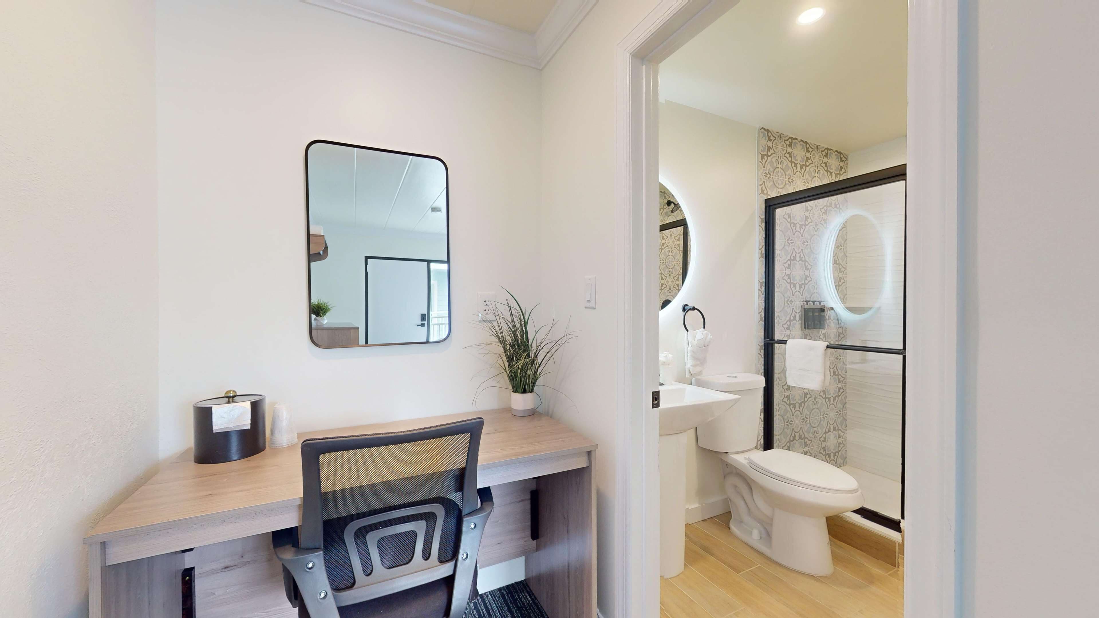 A modern bathroom with a toilet, sink, and mirror. There is a desk with a chair and a potted plant.