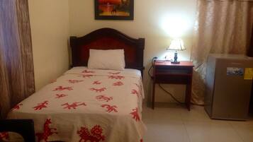 Habitación de hotel con una cama, una mesita de noche con lámpara, un refrigerador y un cuadro en la pared.