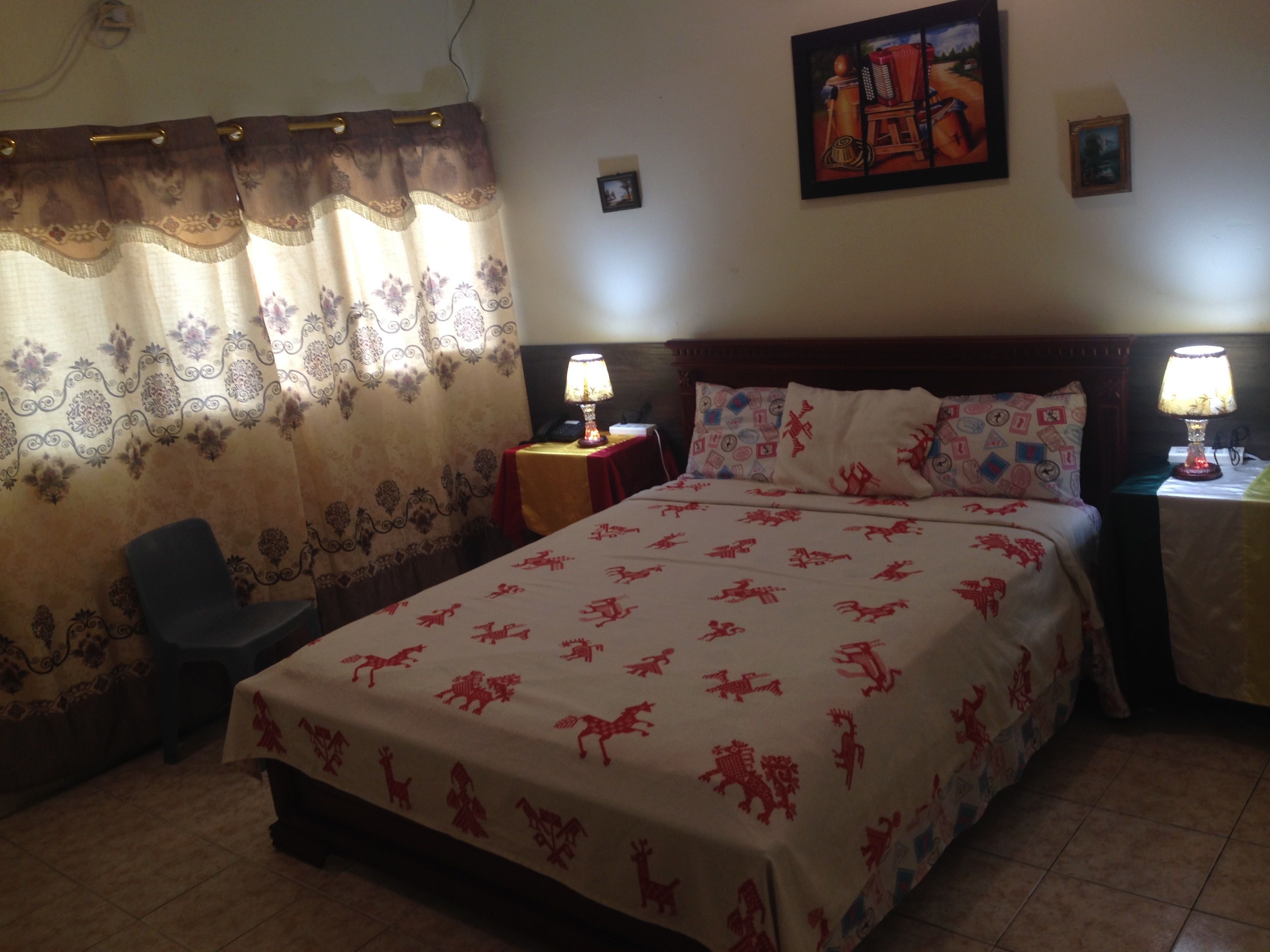 A hotel room with a large bed, two bedside lamps, a chair, a small table, and a curtain with a floral pattern.