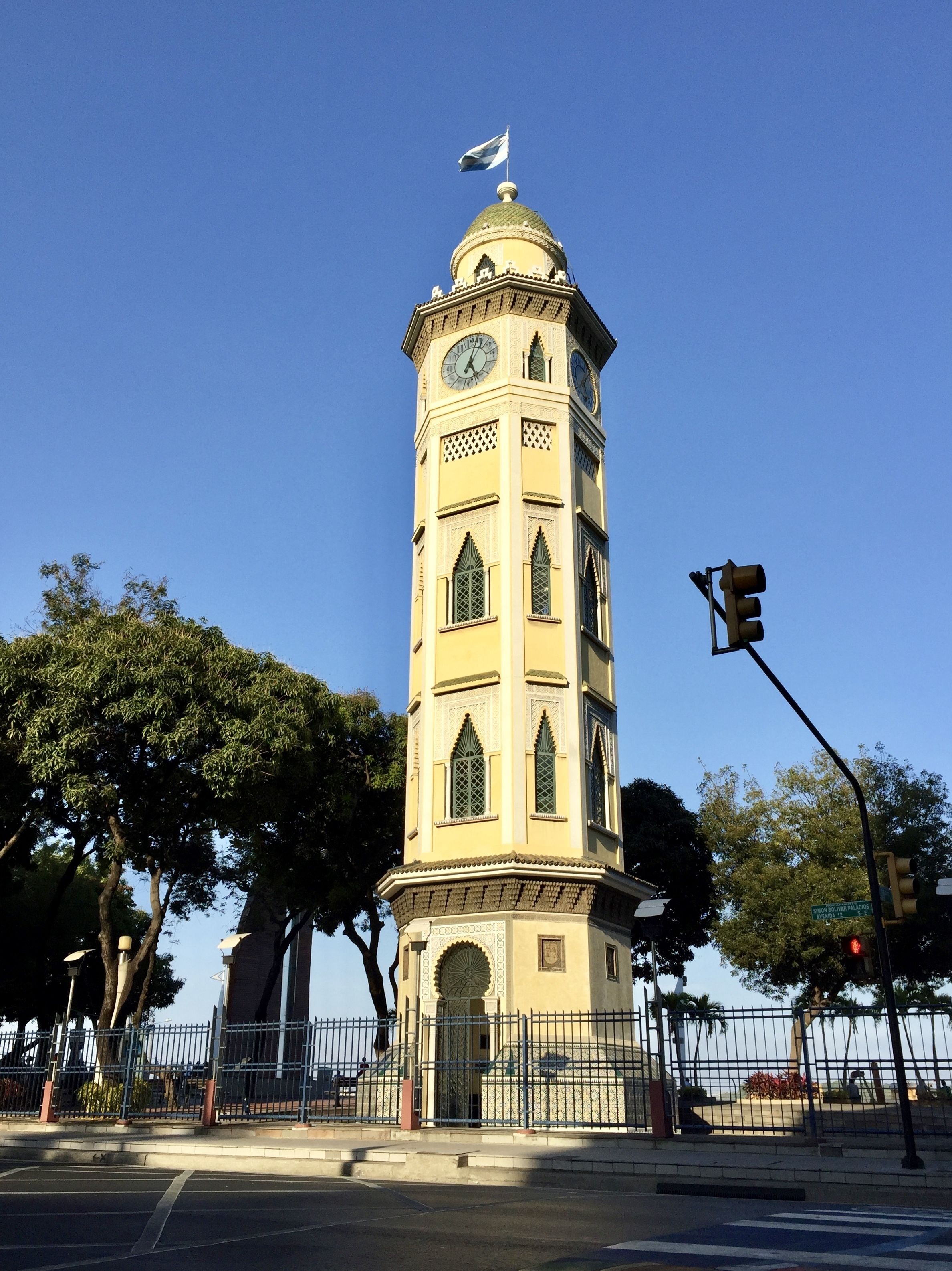 Una torre alta de color beix amb un rellotge i una bandera a la part superior, envoltada d'arbres i una tanca.