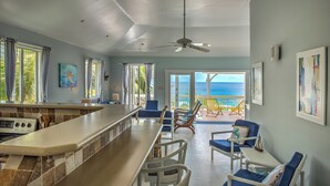 A spacious living area with a kitchen counter, dining table, and chairs. There are large windows with curtains, a ceiling fan, and artwork on the walls. - White Caps (Bathsheba)