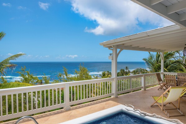 A pool area with a view of the ocean, wooden chairs, and a white railing.