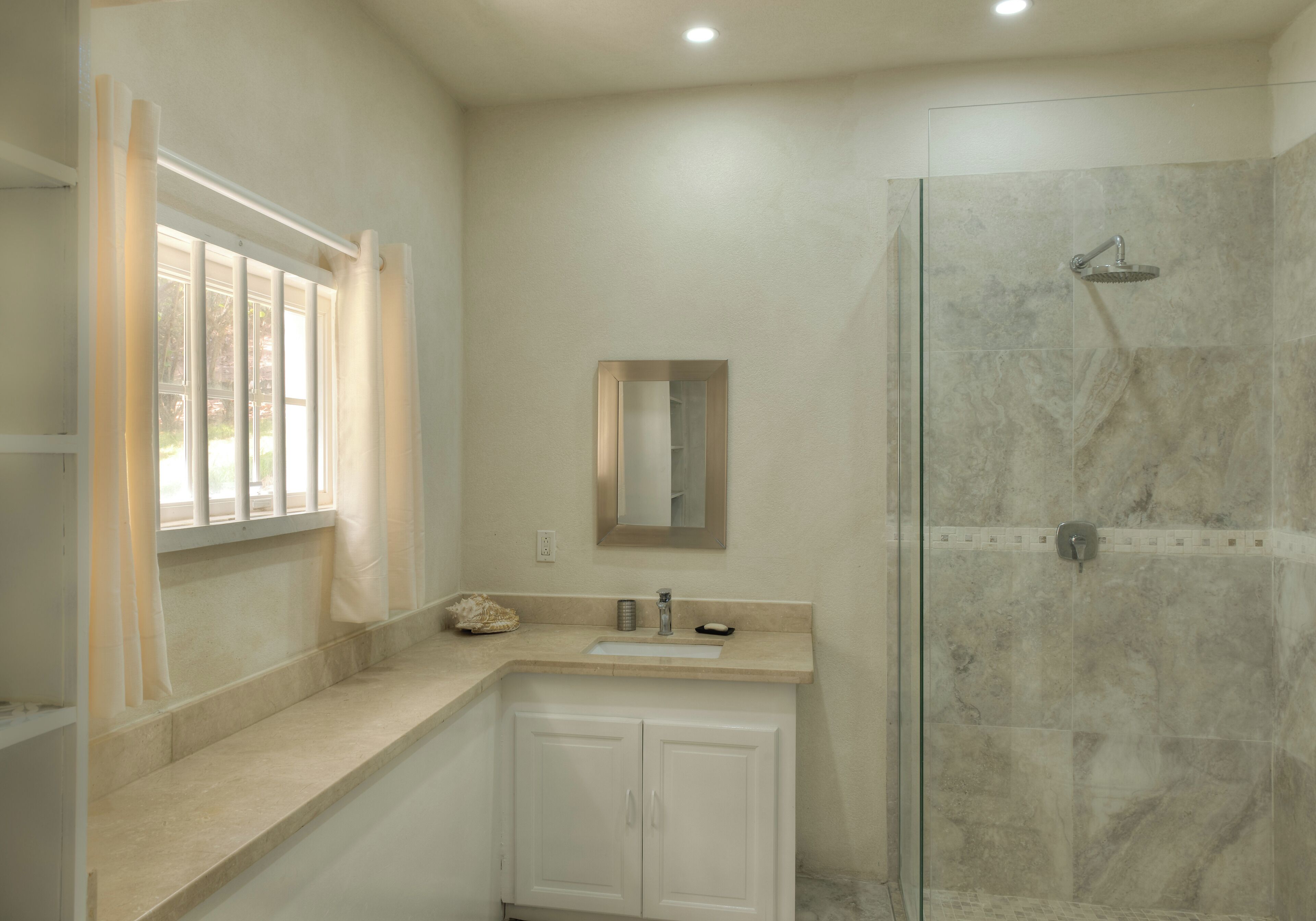 A bathroom with a glass shower enclosure, a sink with a mirror above it, and a window with curtains.