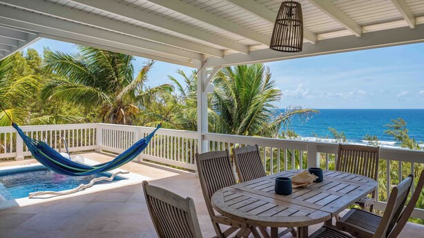 A covered outdoor area with a pool, wooden furniture, and a view of the ocean. - White Caps (Bathsheba)