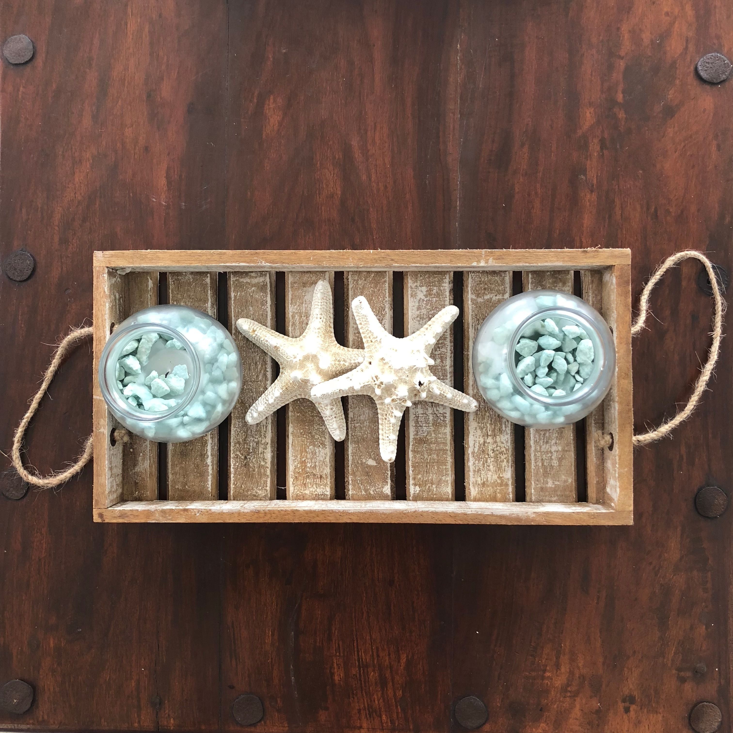 Un plateau en bois contenant deux bocaux en verre remplis de petits cailloux bleus, une étoile de mer blanche et un fond en bois rustique.