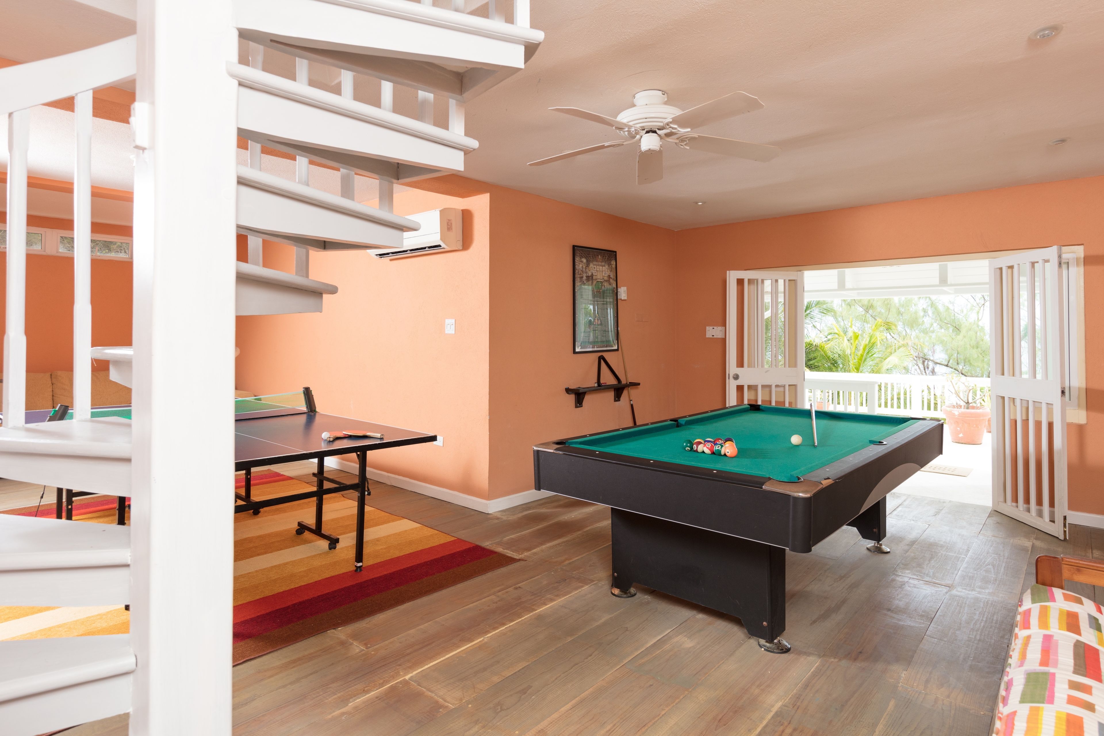 A pool table in a room with orange walls, a ceiling fan, and a view of greenery outside.