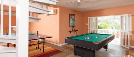A pool table in a room with orange walls, a ceiling fan, and a view of greenery outside.