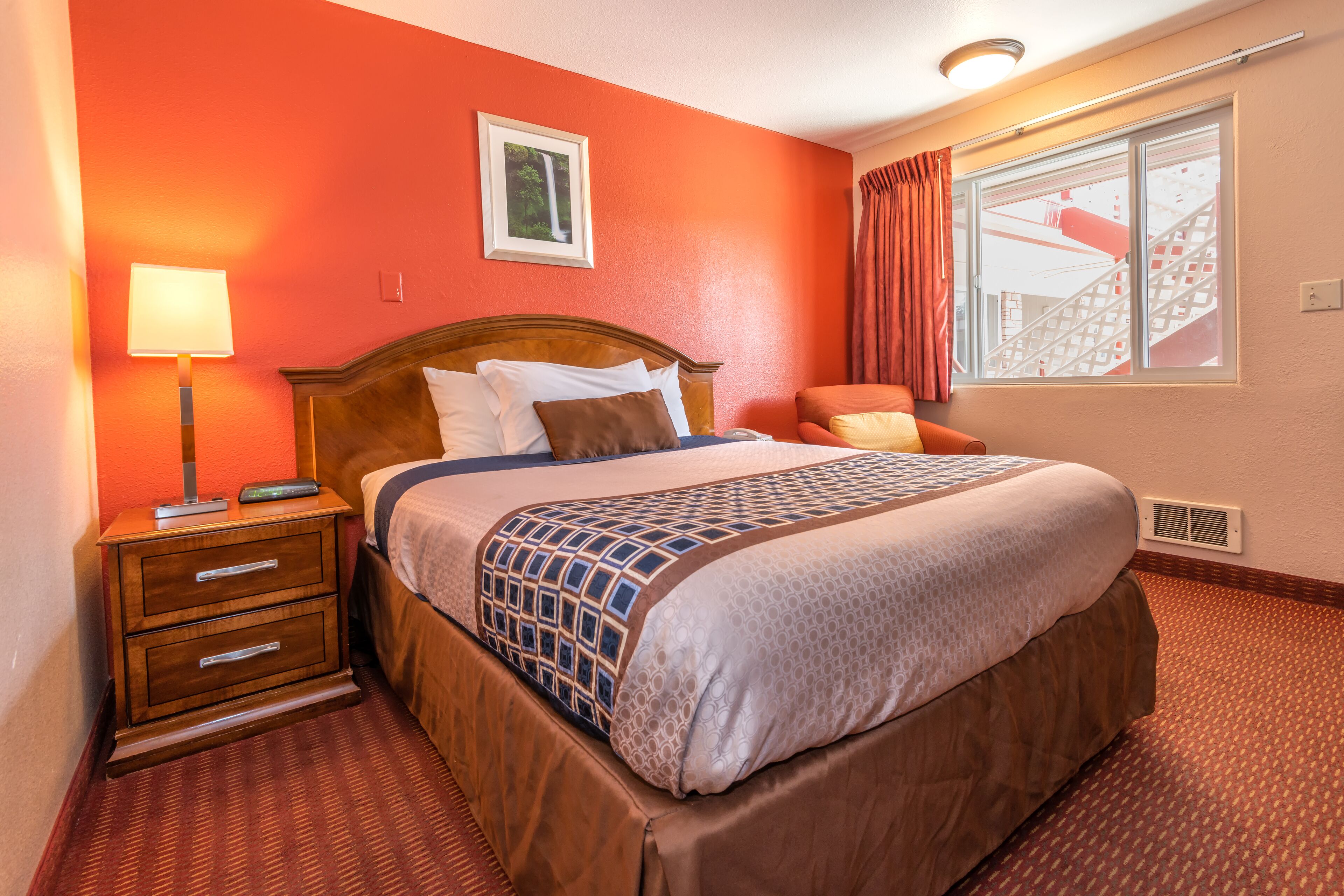 A hotel room with a large bed, a nightstand with drawers, a lamp, a chair, and a window with curtains.