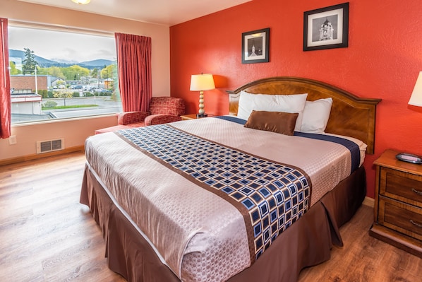 A hotel room with a large bed, a wooden nightstand, a chair, and a window with a view of a parking lot and mountains.