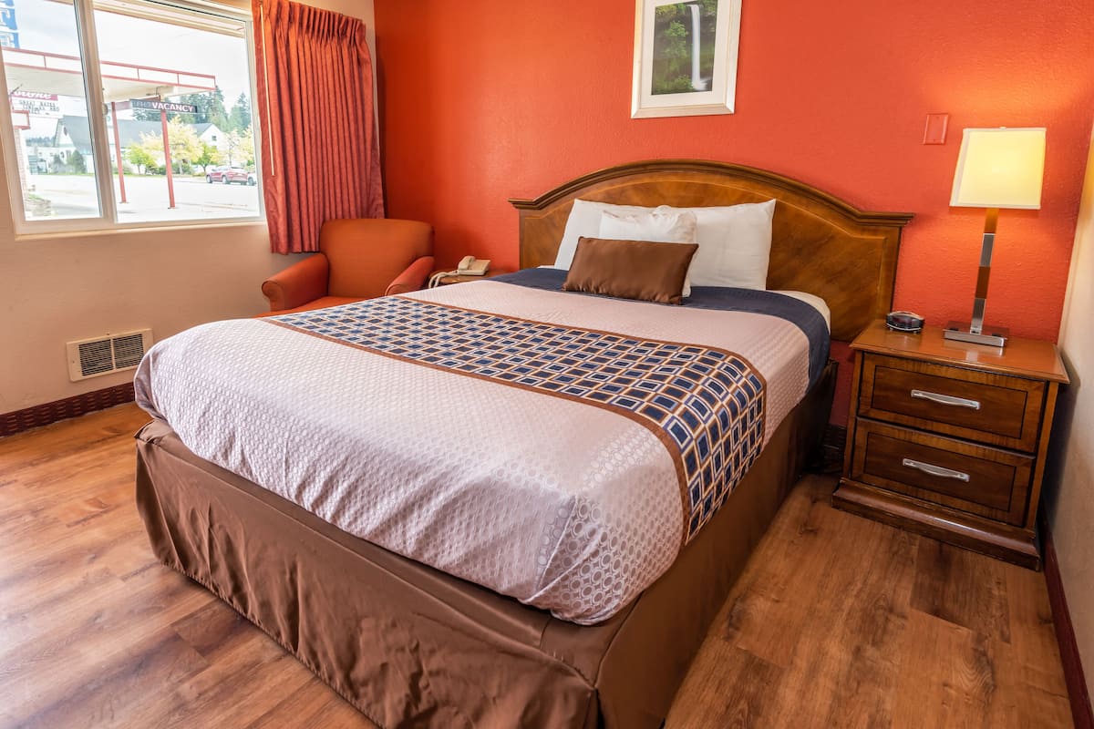 A hotel room with a large bed, a wooden nightstand with drawers, a chair, and a window with a view of the outside.