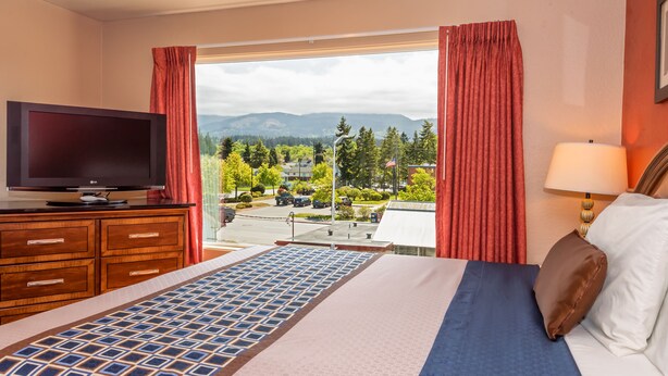 A hotel room with a bed, a TV, a window with curtains, and a view of a parking area and buildings. - Flagstone Motel (Port Angeles)
