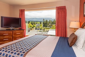 A hotel room with a bed, a TV, a window with curtains, and a view of a parking area and buildings.