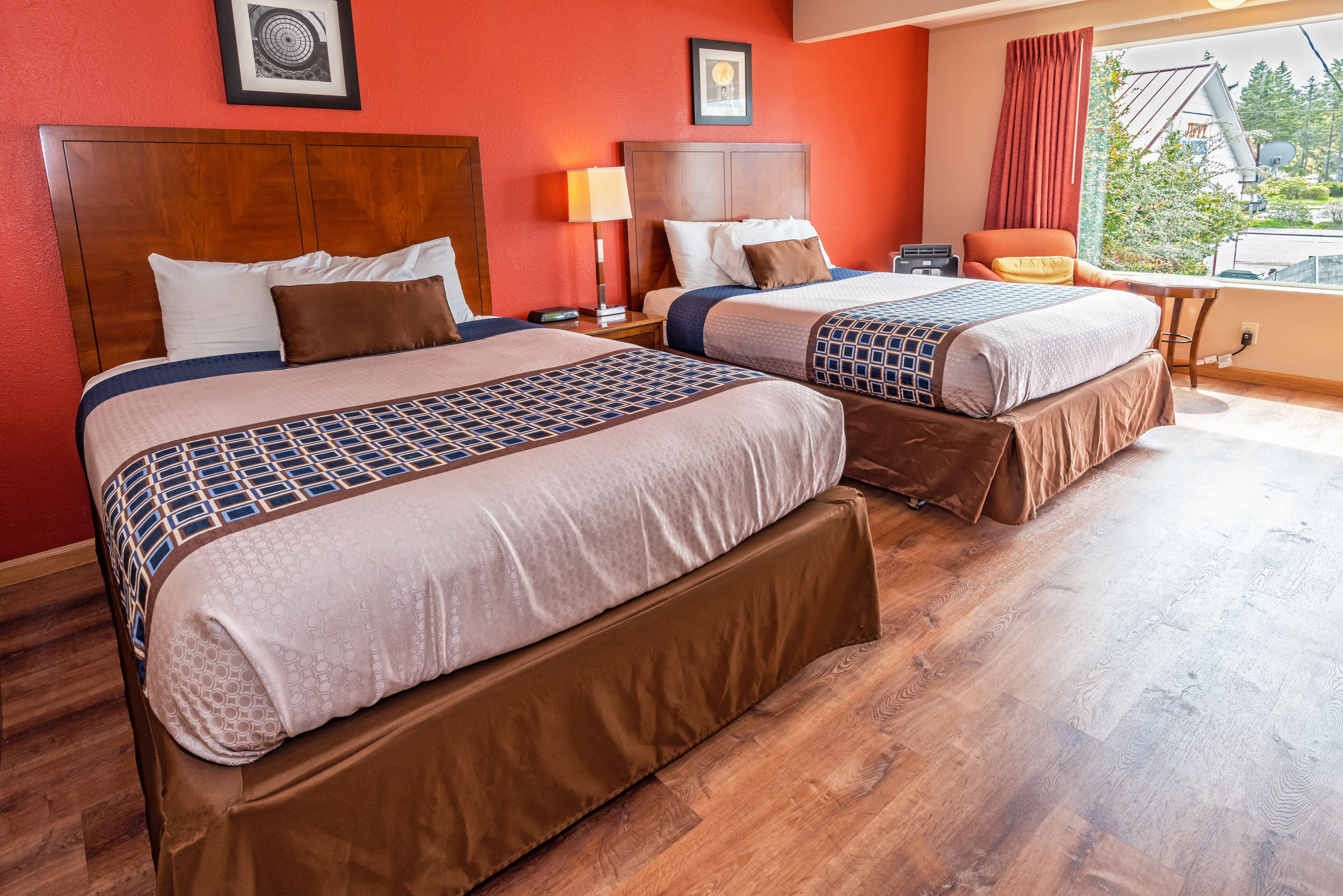 A hotel room with two beds, wooden headboards, a window with a view, a desk, and a chair.