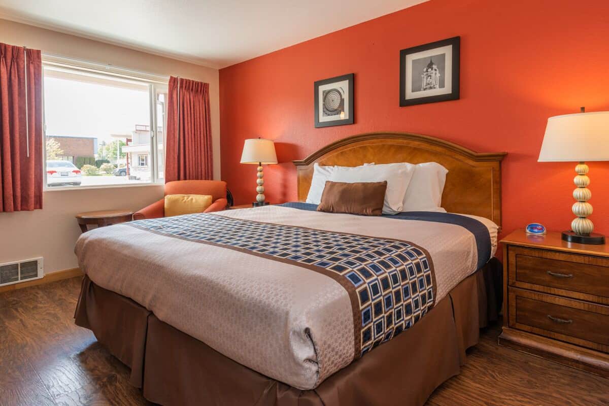 A hotel room with a large bed, a wooden headboard, two bedside tables with lamps, a window with curtains, and two framed pictures on the wall.