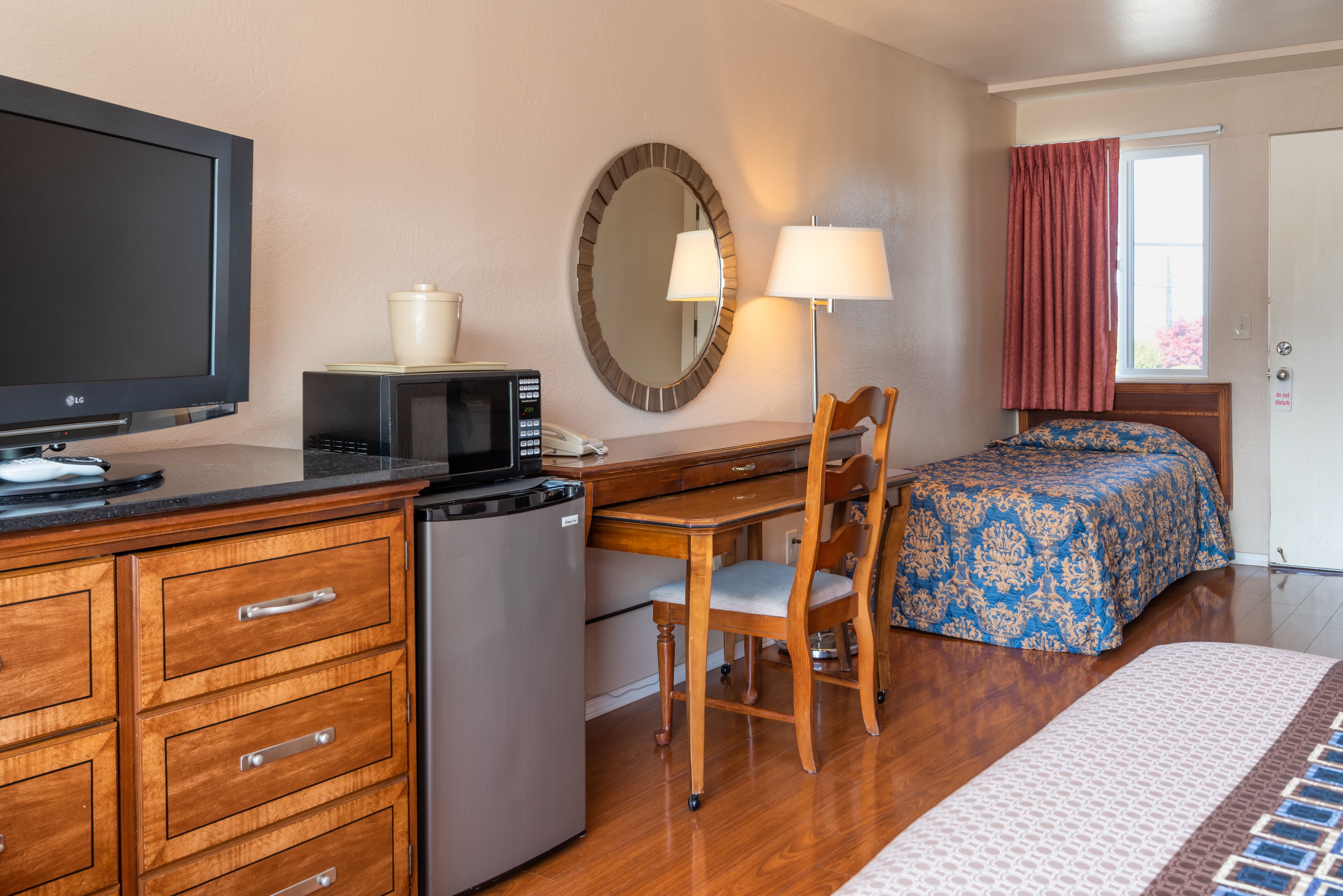 A hotel room with a wooden desk, a bed with a patterned bedspread, a television, a microwave, a refrigerator, a chair, a lamp, and a window with curtains.