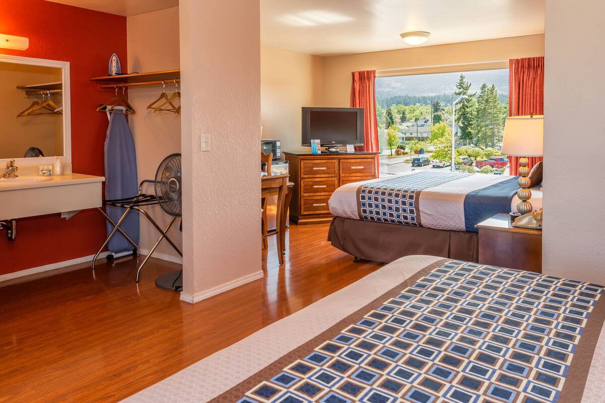 A hotel room with a bed, a desk with a computer, a TV, a window with a view of the outdoors, and a bathroom with a mirror and a sink.