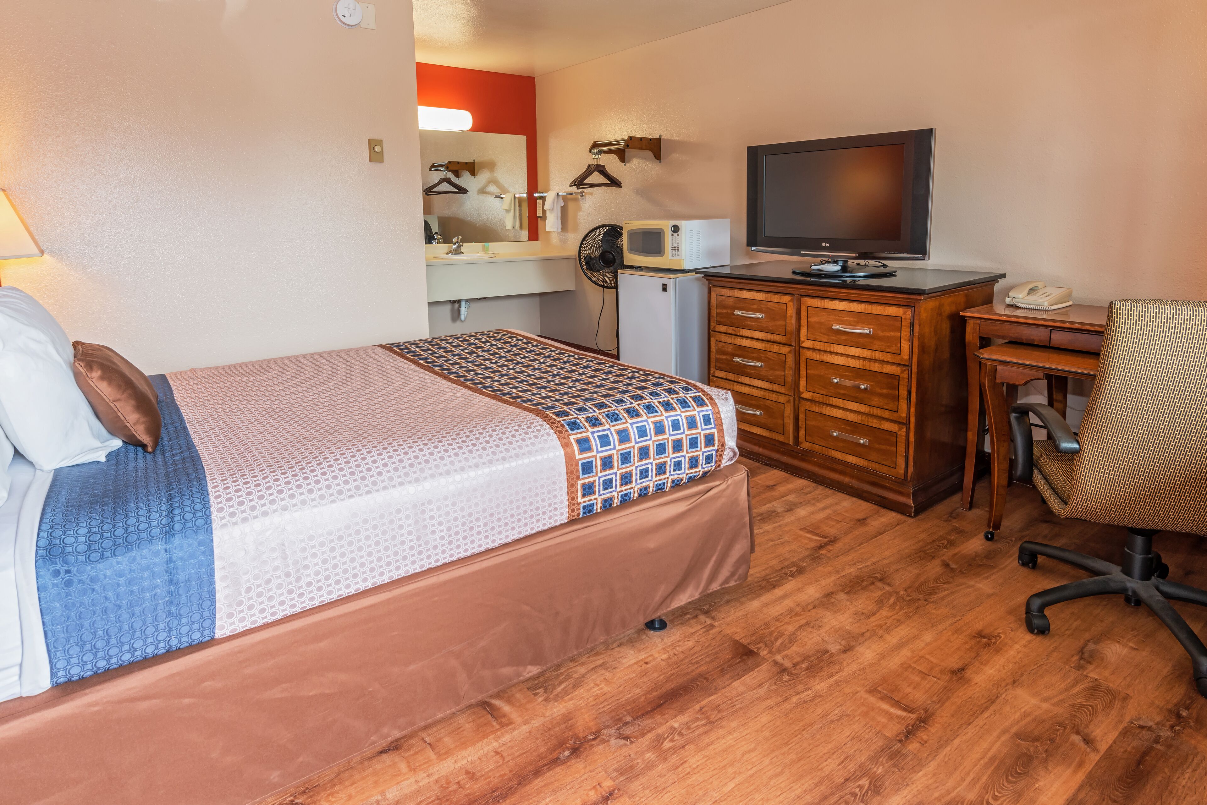 A compact hotel room with a single bed, a wooden desk, a chair, a television, a microwave, and a small refrigerator.