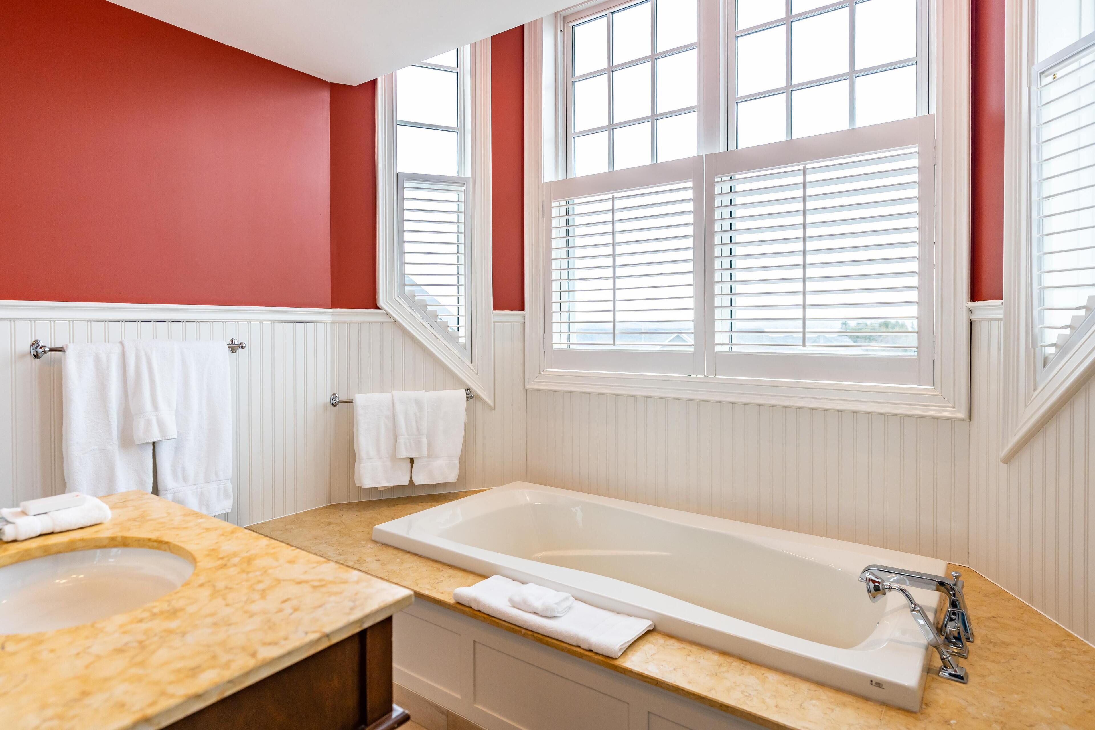 A bathroom with a large bathtub, a sink with a marble countertop, and a window with blinds.