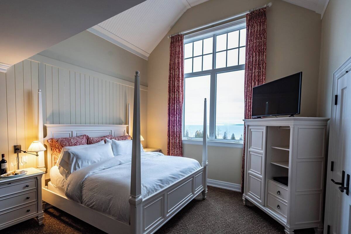 Un lit à baldaquin avec un téléviseur posé sur une commode, une fenêtre avec des rideaux et une table de chevet avec une lampe.