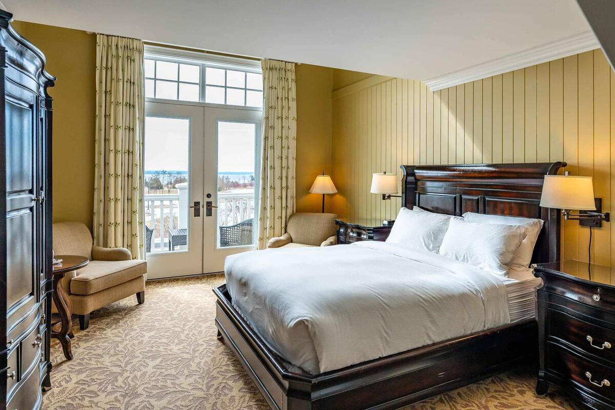 Une chambre d’hôtel avec un grand lit, deux lampes de chevet, une chaise, une commode et une vue sur l’extérieur, visible à travers une porte.
