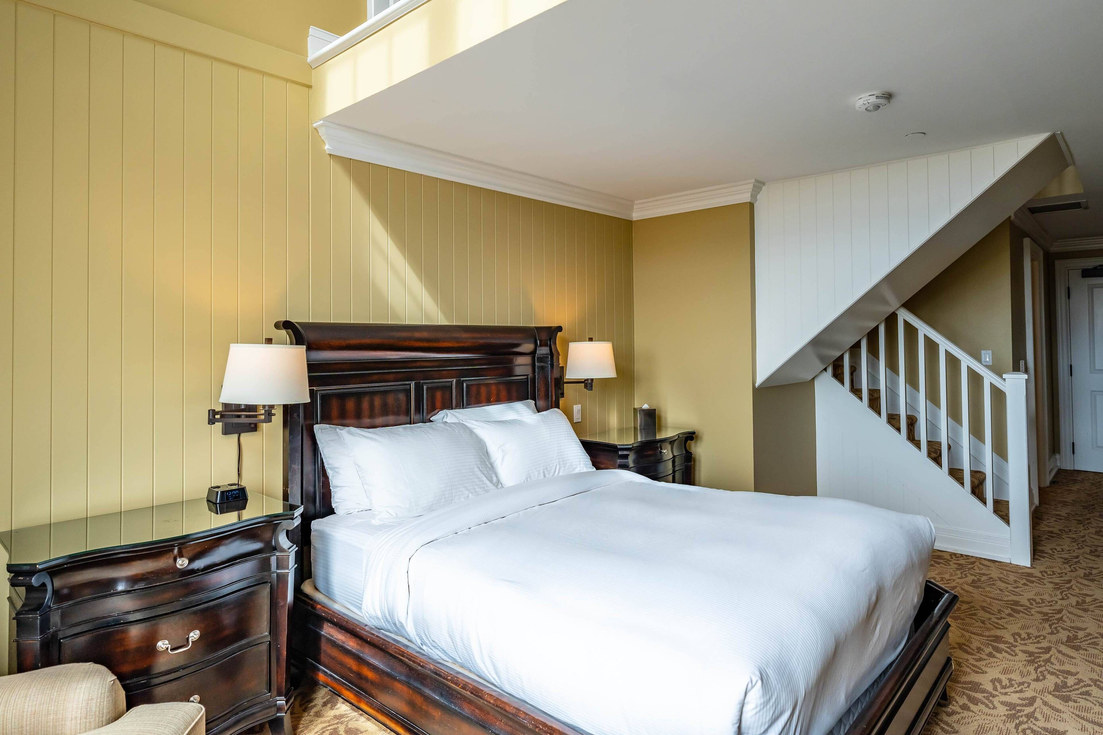 A neatly made bed in a room with a wooden headboard, a nightstand with a lamp, and a staircase to the right.