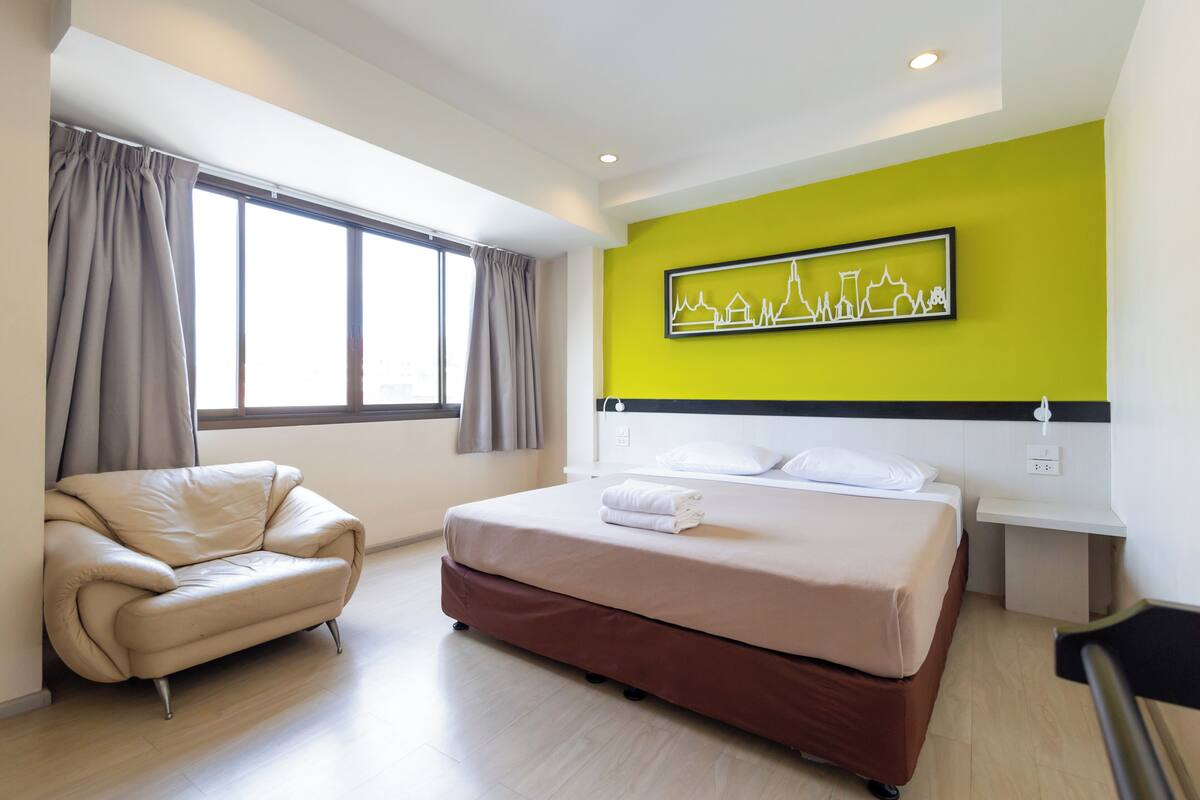 A hotel room with a bed, a sofa, a window with curtains, a wall-mounted artwork, and a bedside table.