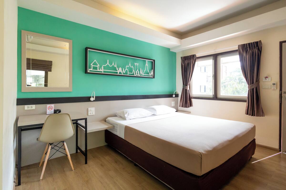 A hotel room with a bed, desk, chair, and a cityscape artwork on the wall.