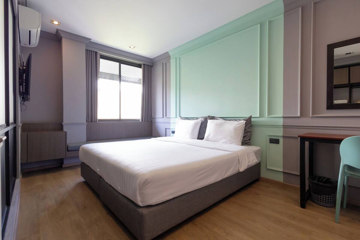 A modern bedroom with a large bed, a window with curtains, a small table, and a chair.