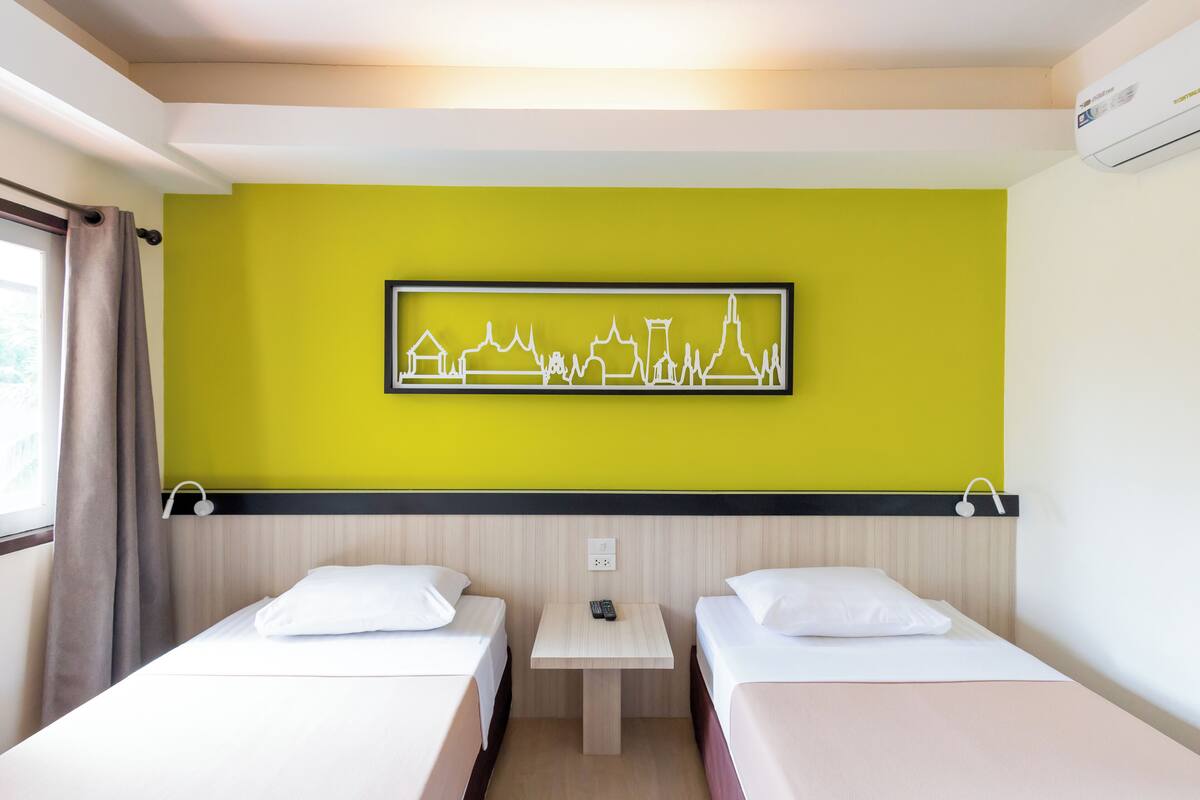 A hotel room with two beds, a small table with a remote control, and a wall-mounted air conditioner.