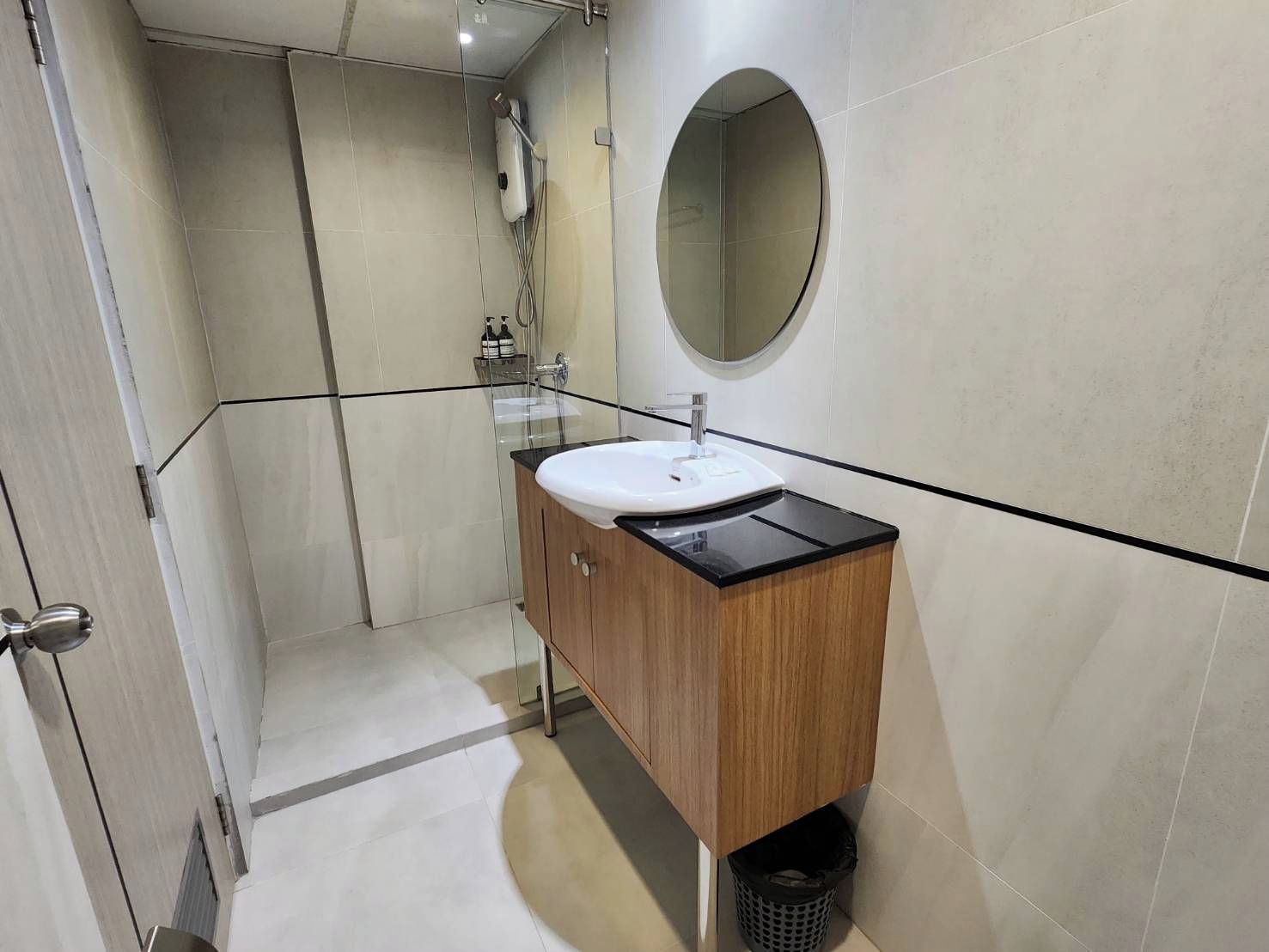 Une salle de bain moderne avec une douche à l’italienne en verre, un meuble-lavabo en bois avec un plan de travail noir et un miroir rond.