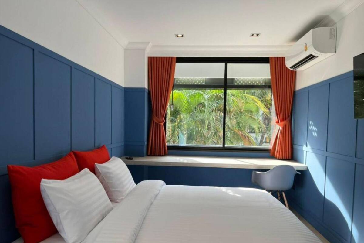 A modern hotel room with a large bed, red and white pillows, a window with curtains, and a view of greenery outside.