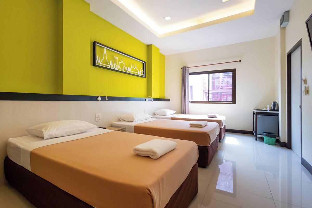A hotel room with three beds, a window with curtains, a wall-mounted artwork, and a small table with a kettle.