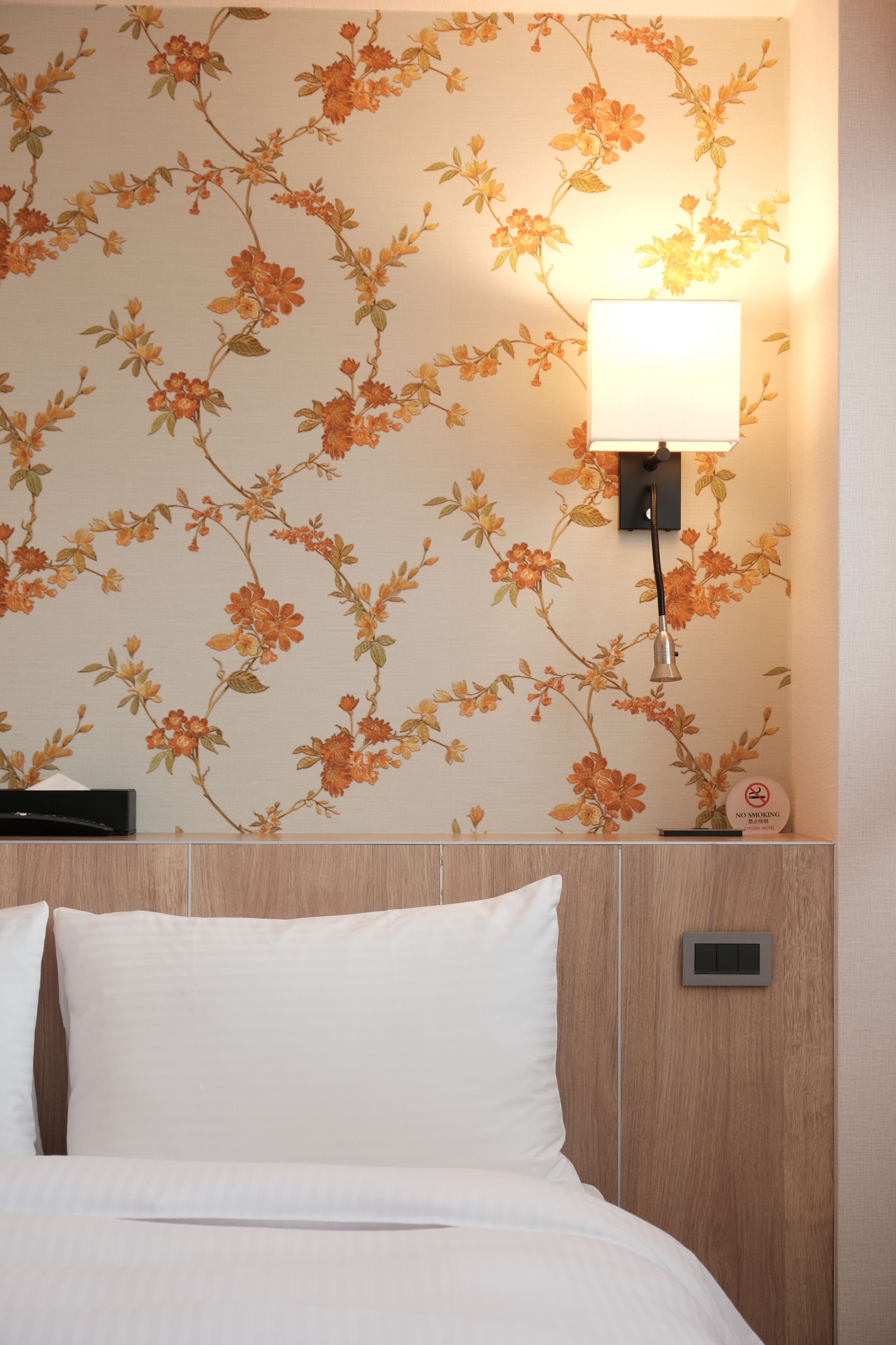 A hotel room with a floral wallpaper, a bedside lamp, a nightstand, and a bed with white bedding.