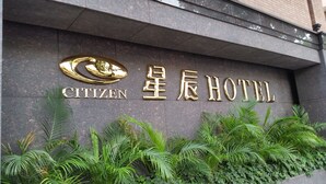Letrero de hotel con el nombre "Citizen" y "星辰 HOTEL" en letras doradas sobre un revestimiento de piedra oscura, rodeado de plantas verdes.