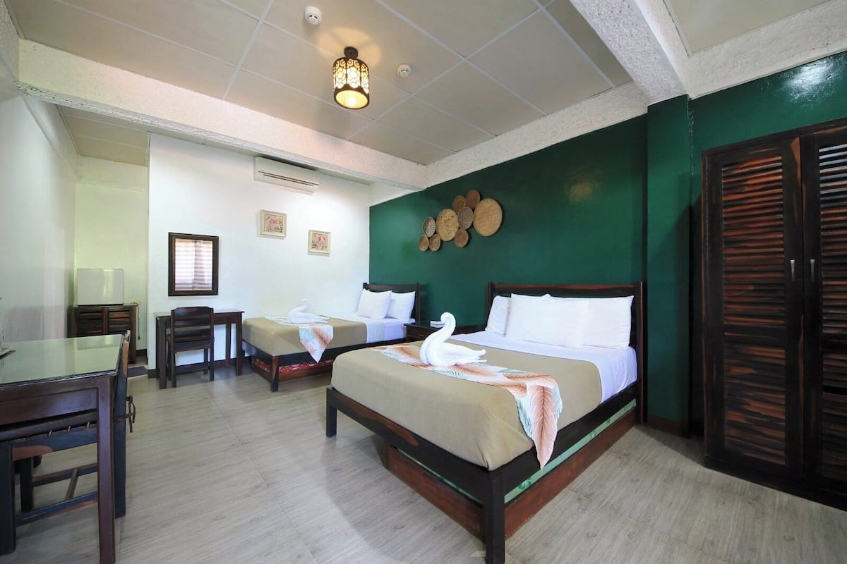A hotel room with two beds, a wooden desk, a chair, a table, a wall-mounted air conditioner, and a ceiling light fixture.