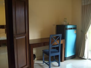 A room with a bed, a small desk with a chair, a blue refrigerator, and a window with curtains.