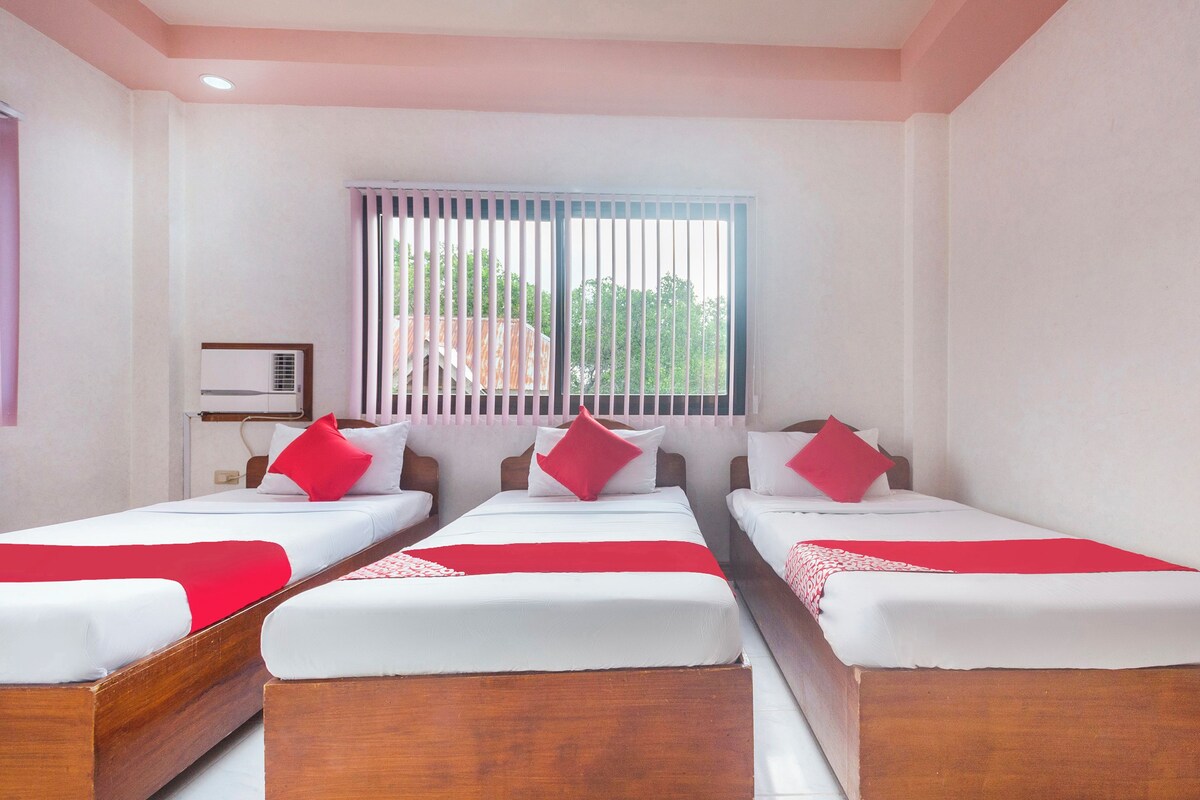 Habitación de hotel con tres camas individuales, cada una con ropa de cama blanca y detalles rojos. Se ve una ventana con persianas verticales, una lámpara de techo y un pequeño equipo de aire acondicionado en la pared.